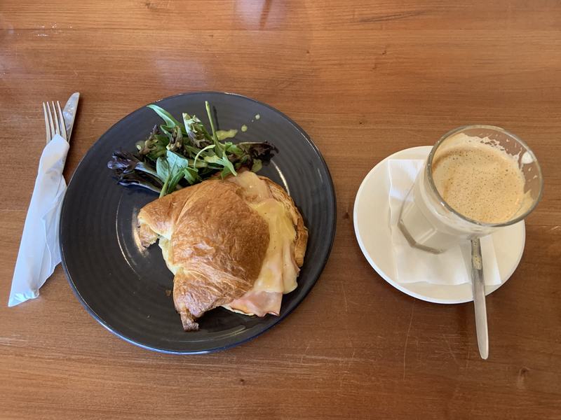 Why it's called Wineglass Bay — A filled croissant with salad on a dark plate, a hot drink beside it, on a woode