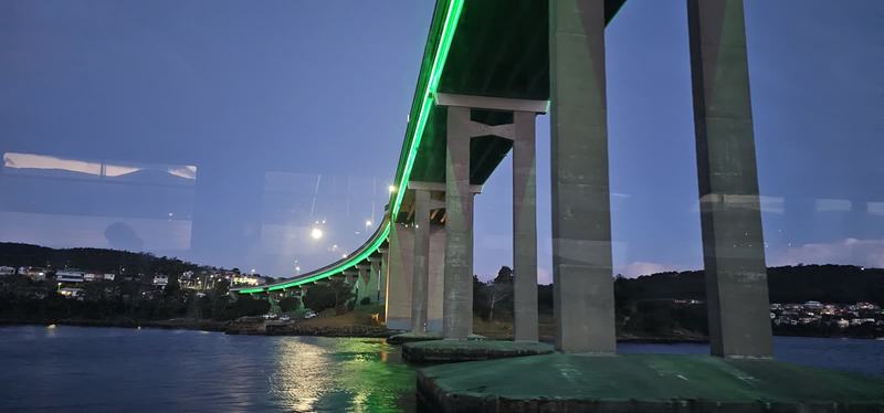 Cut into the cliff β Passing under the Tasman Bridge at dusk: concrete piers and the deck lit green,