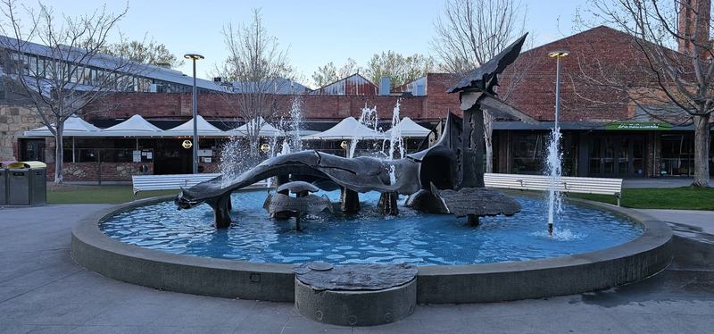 The city that faces south — A bronze fountain in Salamanca Square, water jets around it, brick warehouses be
