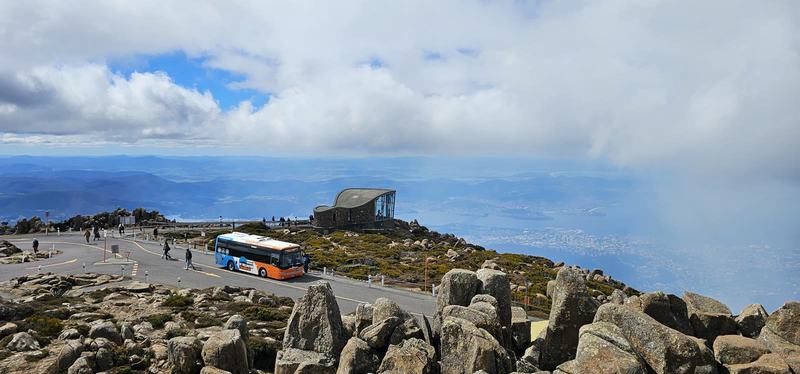 A mountain with four names — The Explorer Bus, orange and blue, parked at the summit loop, the curved stone-a