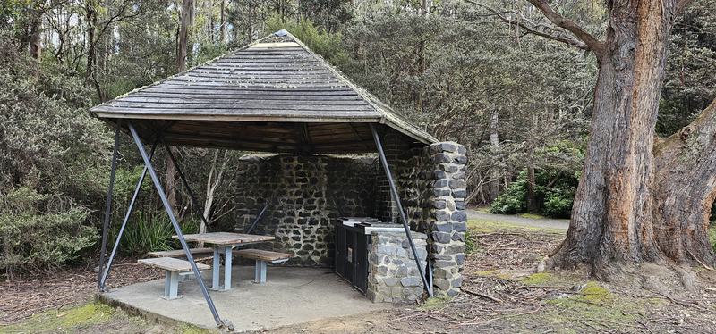 A mountain with four names — A stone-walled picnic shelter with a timber pyramid roof, a table and a barbecue