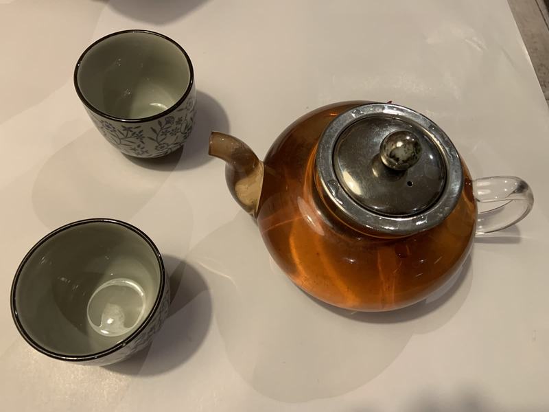 What the gold paid for — A glass teapot of amber tea with a metal lid, beside two small patterned cups