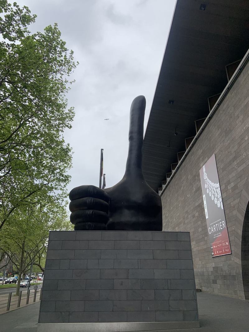 What the gold paid for — A huge black bronze thumbs-up on a stone plinth beside the NGV's stone wall, a C