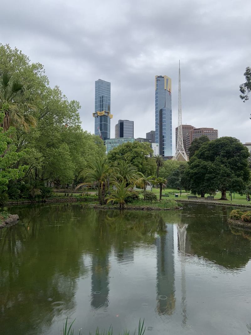 What the gold paid for — A pond reflecting trees and glass towers, with a slim white lattice spire rising